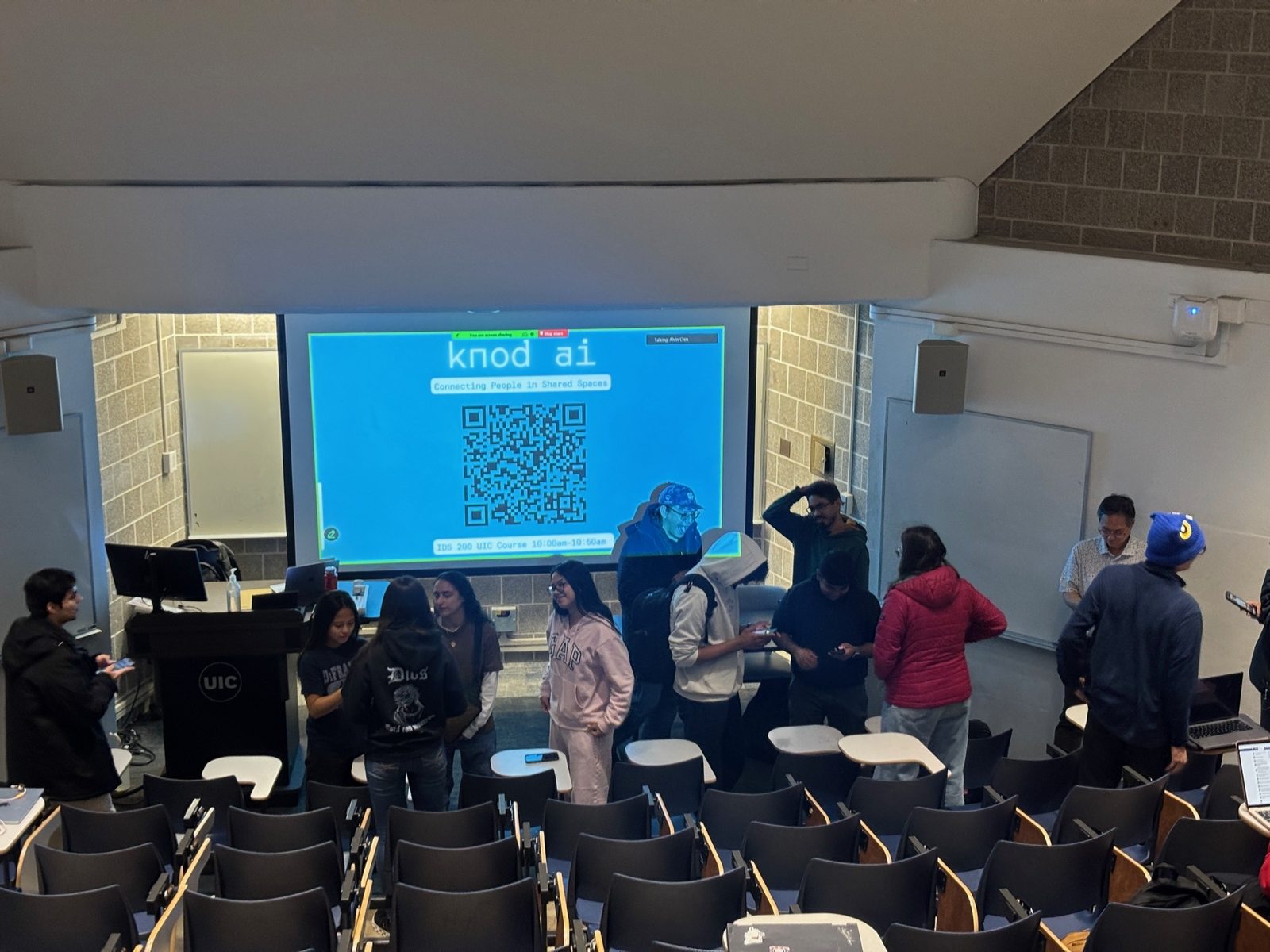 A UIC lecture hall with students mingling and the Knod screen for IDS 200